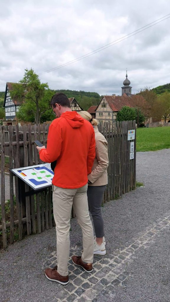 Zwei Personen stehen im Freilichtmuseum vor einem taktil erfahrbaren Übersichtsplan und eine Person hält ein Handy in der Hand. 