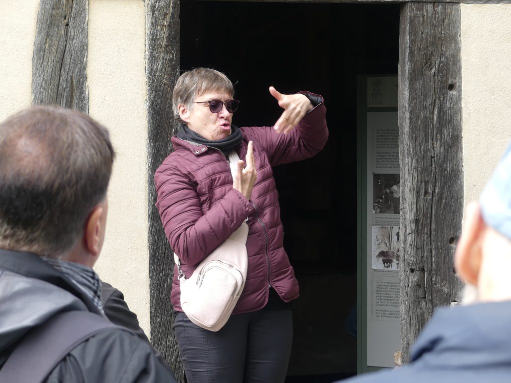 Eine Szene draußen in einem Freilandmuseum. Eine Frau in Jacke und Schal gebärdet im Eingang eines Fachwerkhauses vor einem Publikum. 