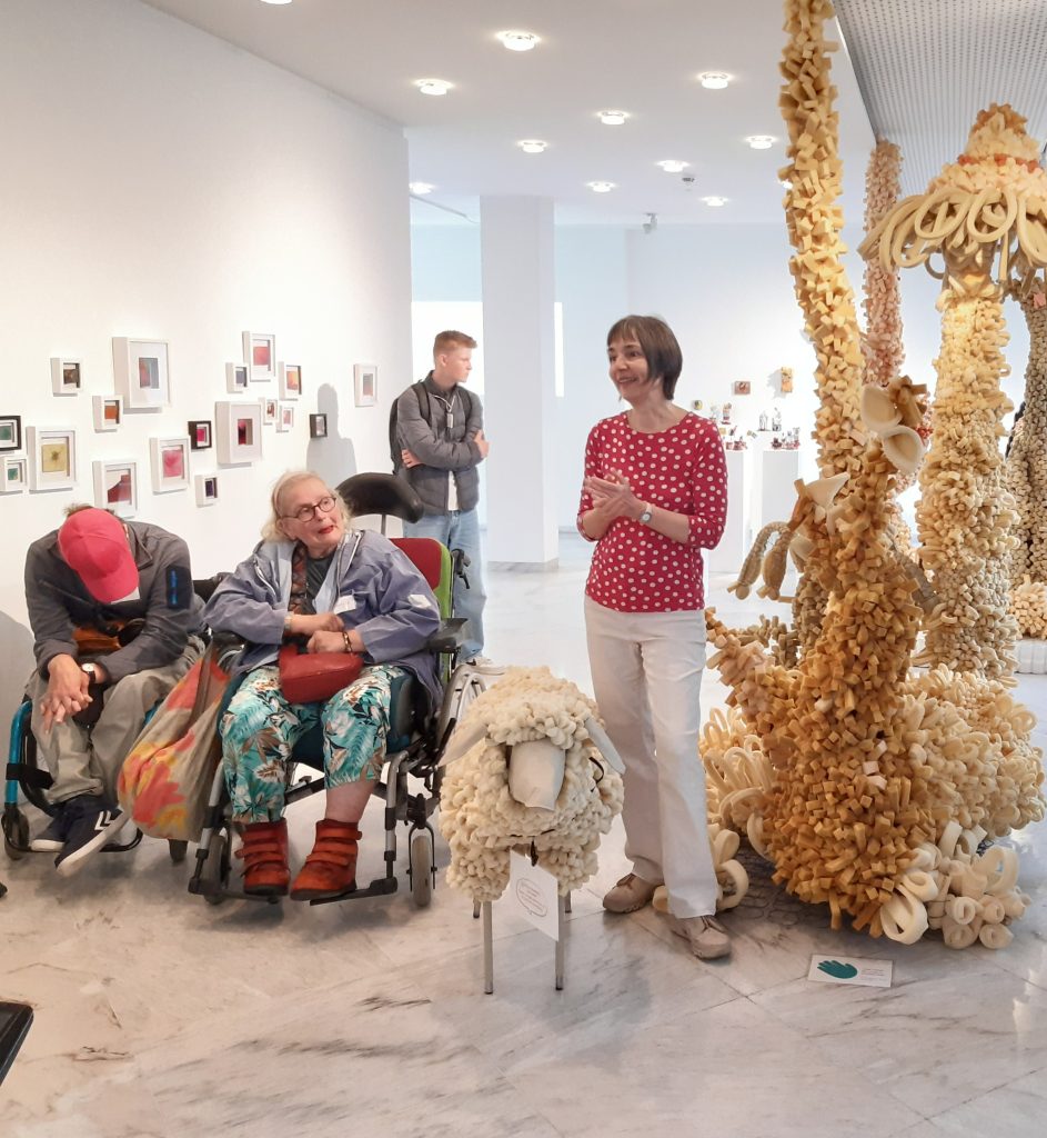 In einer Ausstellung steht Frau Mammel in einem roten Oberteil mit weißen Punkten zwischen Aussstellungsobjekten. Zwei Personen im Rollstuhl sind neben ihr und hören ihr zu. 