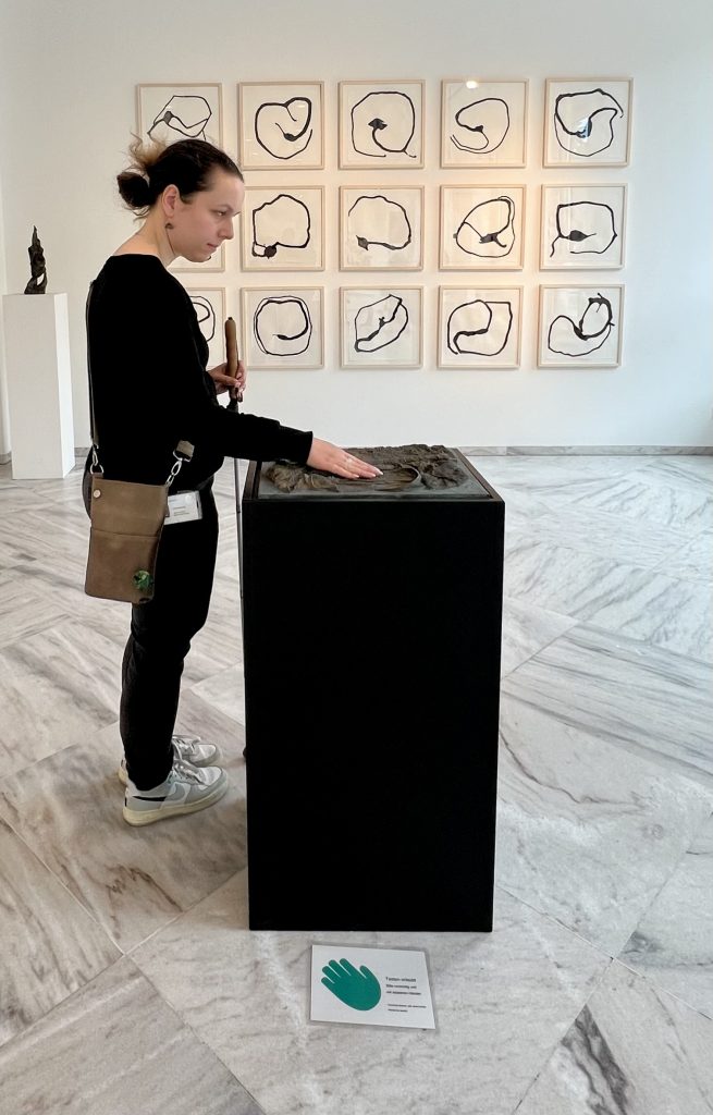 Eine Frau mit Blindenstock steht in einer Ausstellung vor einem hüfthohen Klotz, auf dem ein Relief abgebildet ist. Sie ertastet es mir ihren Händen.