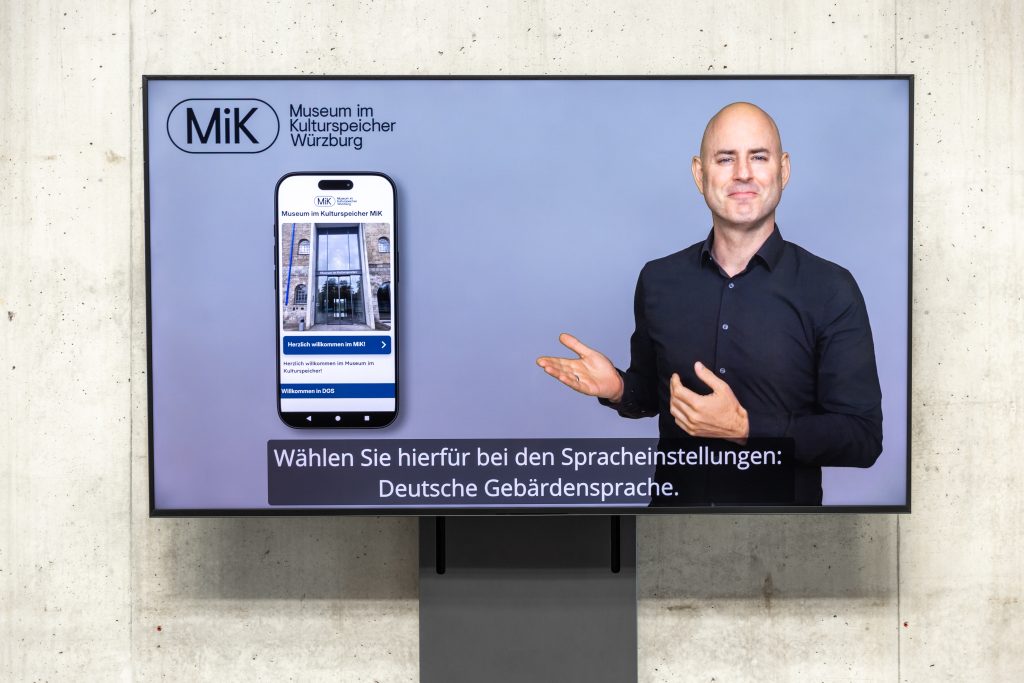 Foto eines Bildschims vor einer Betonwand. Es ist ein Gebärdensprachvideo zu sehen. Ein Mann in schwarzem Hemd gebärdet. Der Screenshot einer Handy-App ist daneben zu sehen. Oben links ist das Logo des MiK zu sehen. 