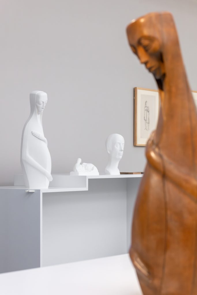Eine Szene in einem Ausstellungsraum. Im Vordergrund ist die Originalskulptur einer Frau aus Holz zu sehen. Im Hintergrund weiße Tastmodelle der Skulptur und zwei weitere Tasmodelle.